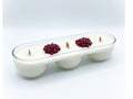 A white three-wick candle in a clear glass elongated container with three small red flower decorations on top, set against a light gray gradient background.
