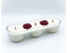 A white three-wick candle in a clear glass elongated container with three small red flower decorations on top, set against a light gray gradient background.