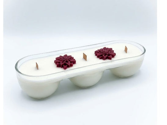 A white three-wick candle in a clear glass elongated container with three small red flower decorations on top, set against a light gray gradient background.