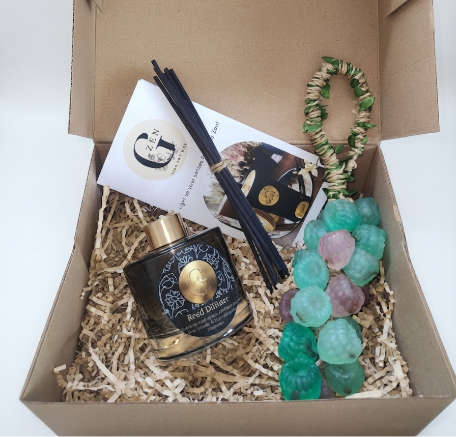 A curated gift box set containing a black and gold reed diffuser bottle, black reeds, a white branded card, a black and gold jewelry pouch, and a green and purple beaded soap charm, all arranged in shredded paper inside an open brown cardboard box.