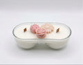 A double-wick soy candle in a clear glass bowl-shaped container, filled with white wax and decorated with two pink flower-shaped ornaments on top.