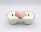 A double-wick soy candle in a clear glass bowl-shaped container, filled with white wax and decorated with two pink flower-shaped ornaments on top.