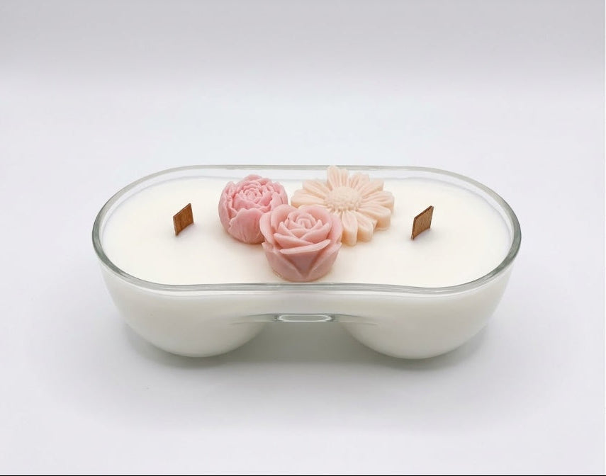 A double-wick soy candle in a clear glass bowl-shaped container, filled with white wax and decorated with two pink flower-shaped ornaments on top.