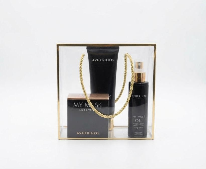 A three-piece  of Avgerinos skincare gift set in a clear acrylic box with a gold frame and rope handle. The set includes a black body cream my musk tube, a perfume musk, and a body musk oil bottle with a gold spray nozzle.