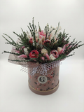 A dried flower arrangement in a round floral box with lace trim, photographed on a plain light background. The bouquet includes pink and cream scented wax roses, white filler flowers, and greenery, with the box printed in a muted pink floral pattern and a circular monogram logo.