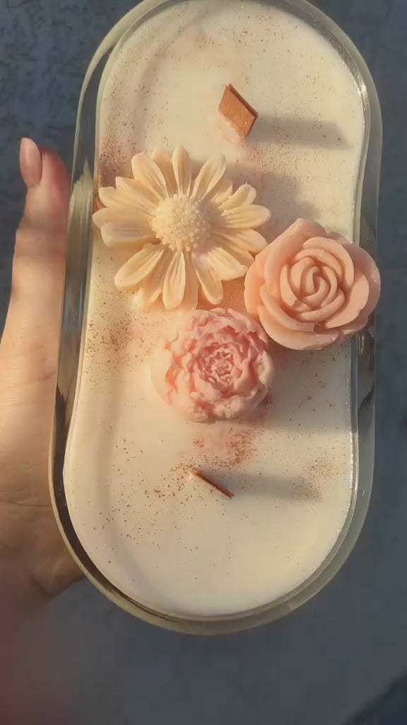 An oval glass jar candle filled with creamy off-white wax, decorated with three sculpted flowers in shades of pink and peach, and topped with a light brown cinnamon stick, shown from a top-down view on a white background.