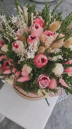 A dried flower arrangement in a round floral box with lace trim, photographed on a plain light background. The bouquet includes pink and cream scented wax roses, white filler flowers, and greenery, with the box printed in a muted pink floral pattern and a circular monogram logo.