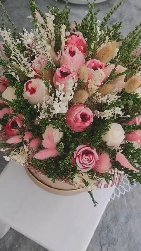 A dried flower arrangement in a round floral box with lace trim, photographed on a plain light background. The bouquet includes pink and cream scented wax roses, white filler flowers, and greenery, with the box printed in a muted pink floral pattern and a circular monogram logo.