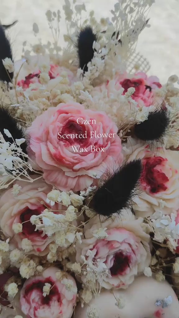 A decorative round black gift box filled with dried flowers, small pink and cream wax roses, black feather-like accents, sheer brown mesh, and a central pale heart ornament, photographed on a light gray studio background. This is a wax flower arrangement handmade by gzen candles base in Cyprus. 