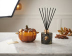 A decorative amber glass pumpkin-shaped diffuser or candle vessel sits beside a dark glass reed diffuser bottle with black reeds on a light marble surface. Small autumn leaves, cinnamon sticks, and a gold card are placed nearby against a softly lit indoor background.