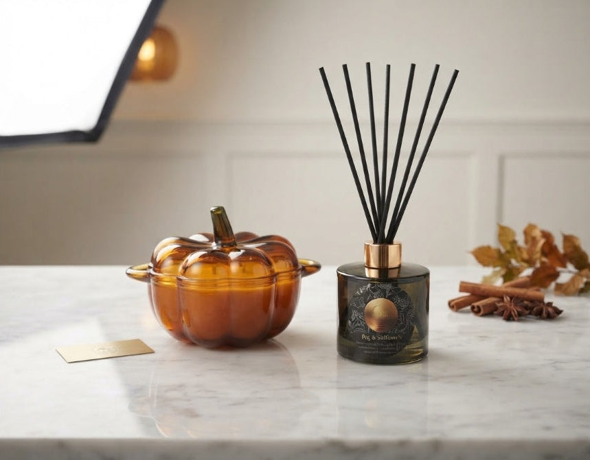 A decorative amber glass pumpkin-shaped diffuser or candle vessel sits beside a dark glass reed diffuser bottle with black reeds on a light marble surface. Small autumn leaves, cinnamon sticks, and a gold card are placed nearby against a softly lit indoor background.
