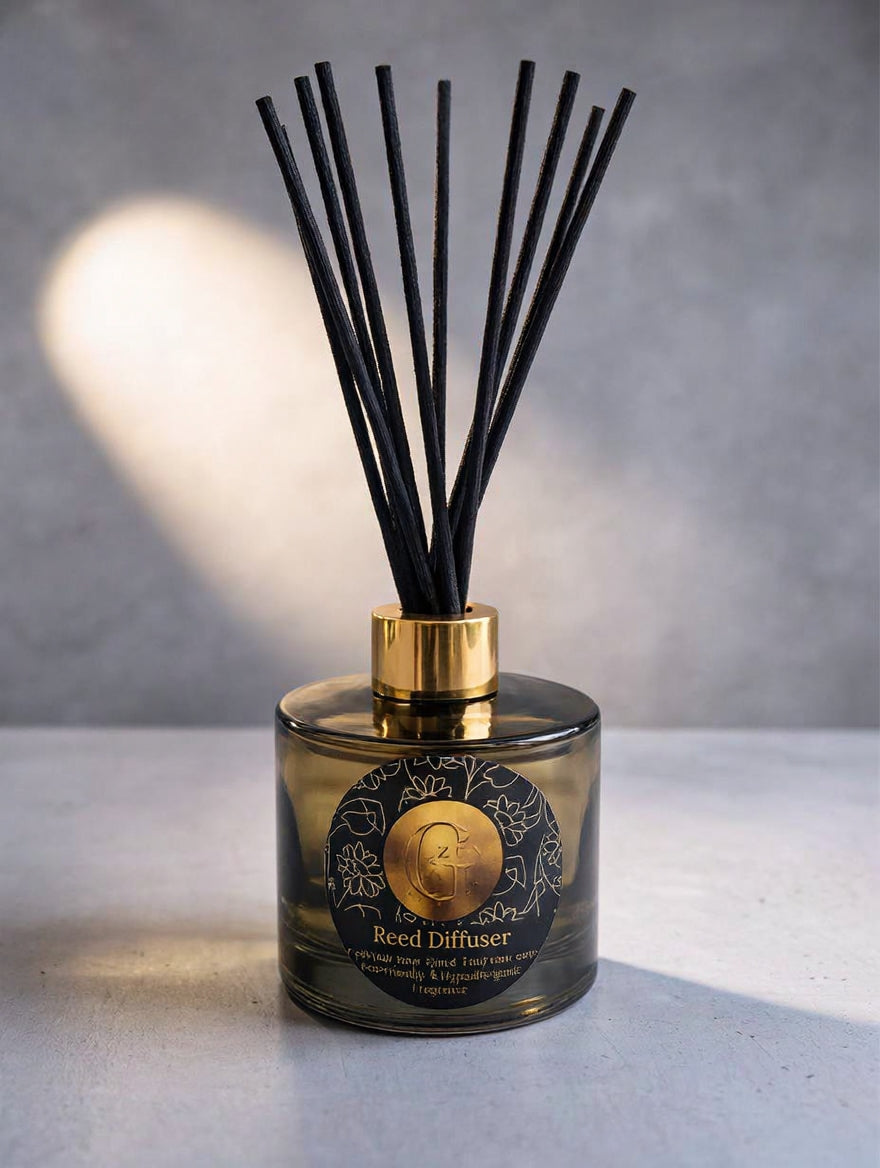 A reed diffuser bottle made of translucent amber glass with a shiny gold collar and black reeds, set against a textured gray background with a soft light reflection.