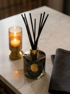 A reed diffuser bottle with black reeds and a gold-toned cap sits on a light marble surface beside a lit candle in a gold holder and a folded dark gray cloth.