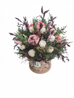 A round floral arrangement featuring pink and white roses, green foliage, and dark burgundy leaves, presented in a decorative round box with a floral pattern, set against a plain white background.