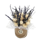A decorative floral candle gift box arrangement in a round kraft paper hatbox with a silver monogram 'G' and pearl trim. The bouquet features cream-colored roses, dark purple lavender sprigs, and white lace accents, set against a plain white background.