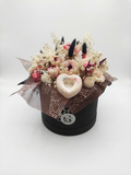 A decorative round black gift box filled with dried flowers, small pink and cream wax roses, black feather-like accents, sheer brown mesh, and a central pale heart ornament, photographed on a light gray studio background. This is a wax flower arrangement handmade by gzen candles base in Cyprus. 