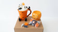 Halloween-themed mug and pumpkin candle set displayed on a brown cardboard box against a white background. The set includes an orange Tigger-shaped mug filled with whipped cream, black polka dots, ghost decorations, and a small pumpkin candle, alongside a jack-o'-lantern-shaped orange candle and small packaged treats.
