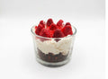 A layered cheesecake candle dessert in a clear glass cup, featuring a dark chocolate crumb base, white cream, and fresh red strawberries on top, set against a plain white background.