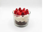 A layered cheesecake candle dessert in a clear glass cup, featuring a dark chocolate crumb base, white cream, and fresh red strawberries on top, set against a plain white background.