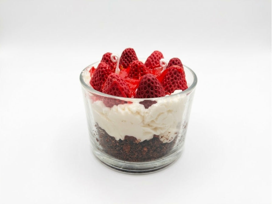 A layered cheesecake candle dessert in a clear glass cup, featuring a dark chocolate crumb base, white cream, and fresh red strawberries on top, set against a plain white background.
