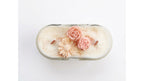 An oval glass jar candle filled with creamy off-white wax, decorated with three sculpted flowers in shades of pink and peach, and topped with a light brown cinnamon stick, shown from a top-down view on a white background.