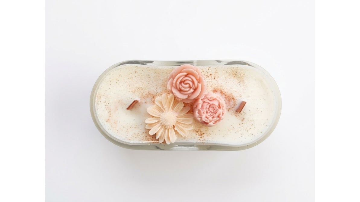 An oval glass jar candle filled with creamy off-white wax, decorated with three sculpted flowers in shades of pink and peach, and topped with a light brown cinnamon stick, shown from a top-down view on a white background.