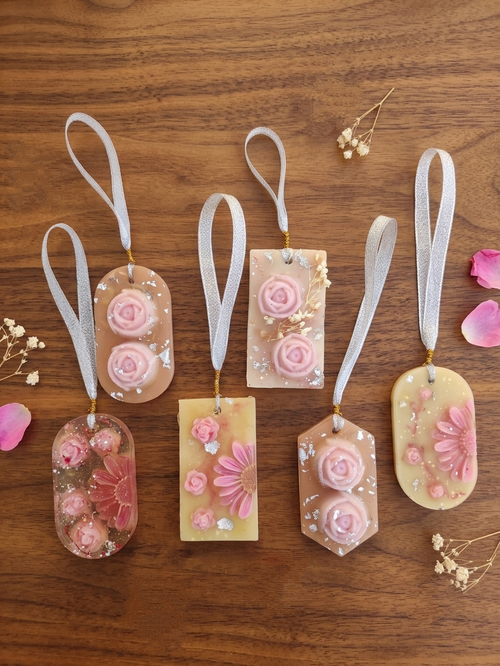 Six decorative scented wardrobe vanilla  keepsake tags with white ribbon loops are arranged on a wooden surface. The tags are translucent and decorated with pink roses, daisies, and glitter, with small dried flowers and pink petals scattered around them.
