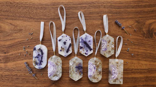 Seven wardrobe handmade soap pendants with hanging loops, decorated with purple lavender flowers and dried botanicals, arranged on a wooden surface with scattered dried lavender sprigs.