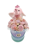 A decorative wax floral gift box arrangement featuring a pink plush elephant wearing a small crown, rising from a pastel lavender and mint round box filled with pink and lavender roses, lace trim, and small beige filler flowers, on a plain white background.
