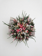 A dried flower arrangement in a round floral box with lace trim, photographed on a plain light background. The bouquet includes pink and cream scented wax roses, white filler flowers, and greenery, with the box printed in a muted pink floral pattern and a circular monogram logo.