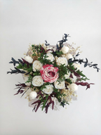 An artificial floral arrangement featuring a central pink soy wax rose surrounded by white wax roses, green foliage, and dark burgundy leaves, set against a plain white background.
