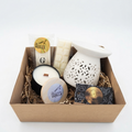 A curated gift box containing a white ceramic diffuser, a white candle in a black tin, a round jar with a vanilla label, a bar of soap, and a packaged item, all arranged in a brown cardboard box with shredded paper on a white background.