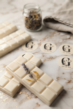 Two cream-colored wax melts bars with a segmented chocolate bar design, decorated with dried lavender and gold leaf flakes, arranged on a white marble surface. Several round paper labels with the brand 'ZEN' are scattered around, along with a small jar of dried botanicals and a folded linen cloth in the background.
