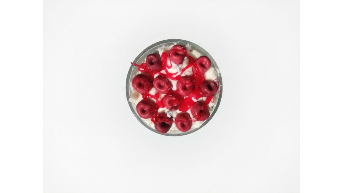 A top-down view of a clear glass bowl filled with white whipped candle  cream and topped with red candle  cherries and red syrup drizzle, set against a plain white background.
